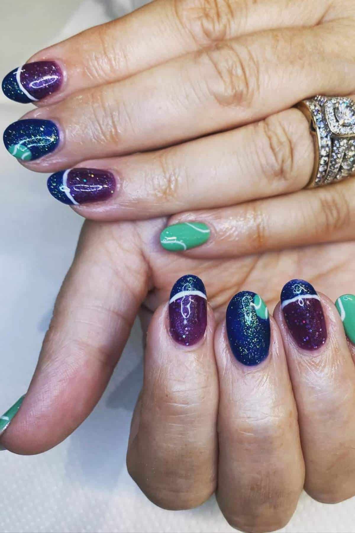 Close-up of hands with manicured nails featuring a mix of purple with glitter, green, and blue wavy patterns; a diamond ring is worn on one finger.