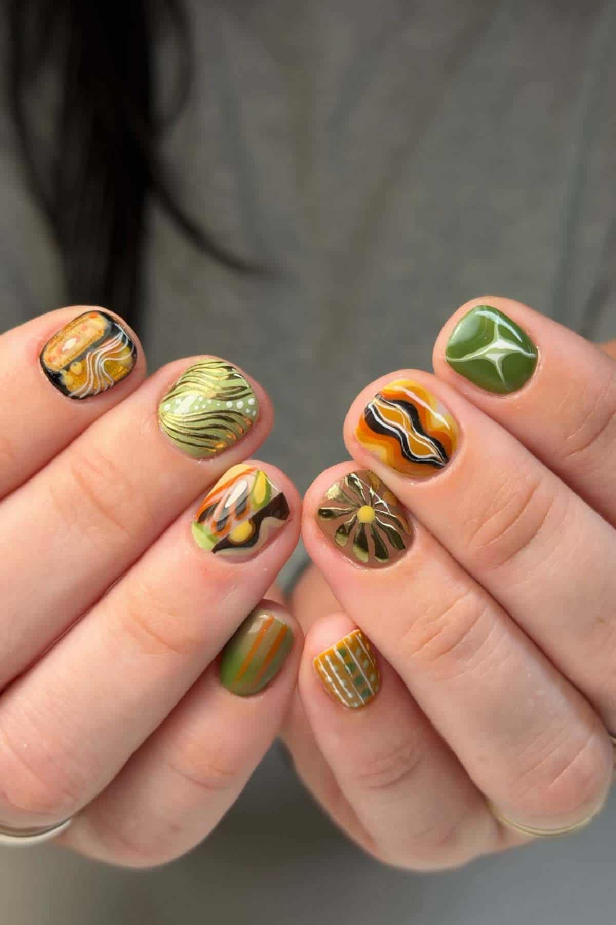 Close-up of hands with short fingernails featuring various green, yellow, orange, and black nail art designs, including stripes, swirls, and geometric patterns.