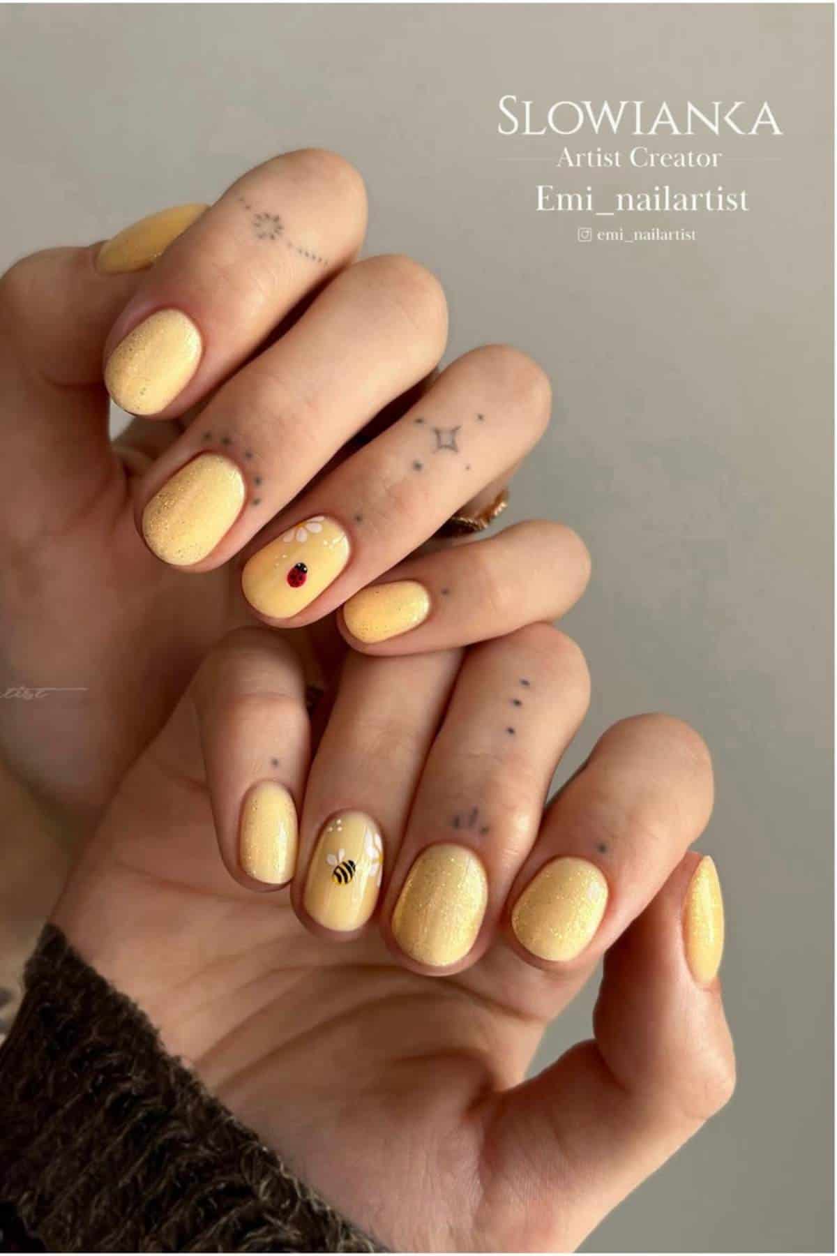 A hand with short, yellow, shimmery nails, featuring a bee design and a red gem accent; visible small tattoos on the fingers and hands.