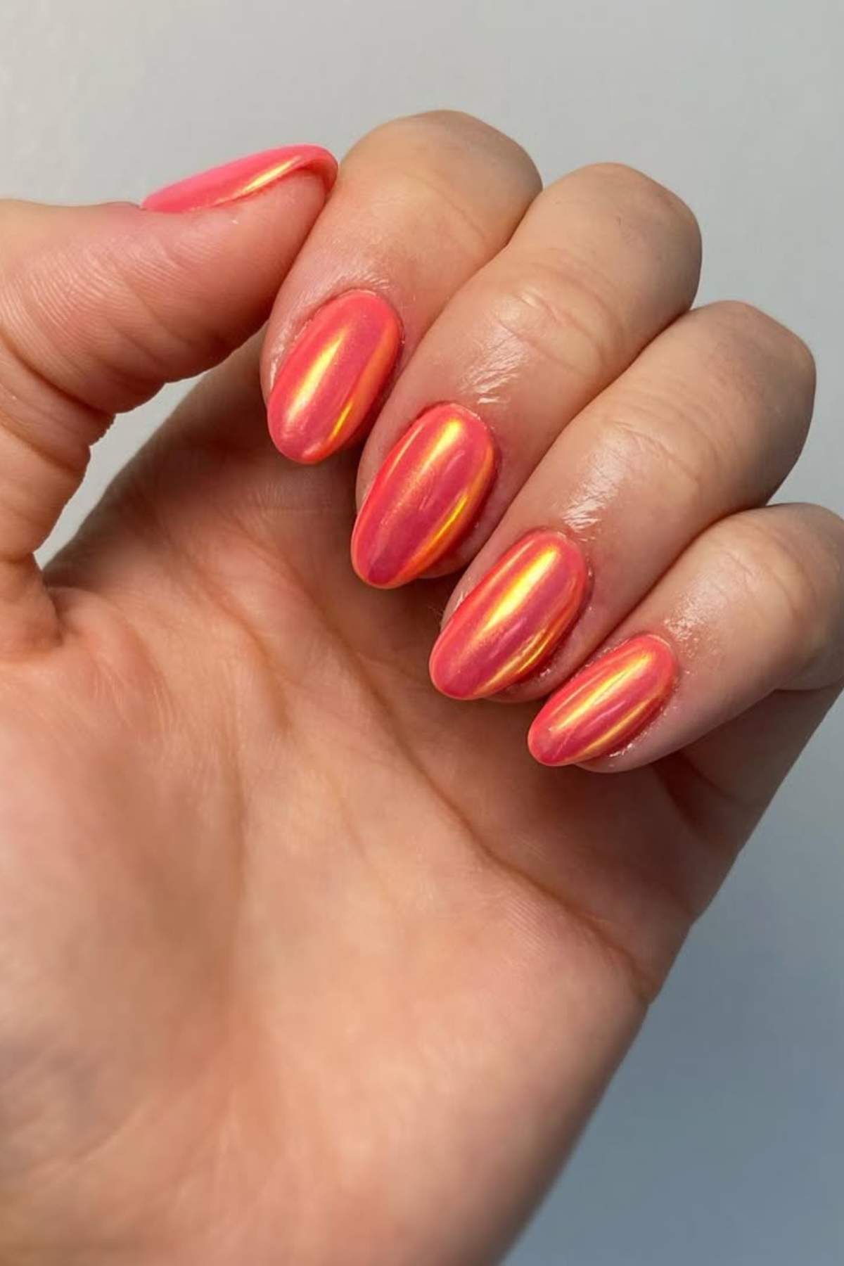 A hand with short, oval-shaped nails painted in a shiny, iridescent coral pink polish.