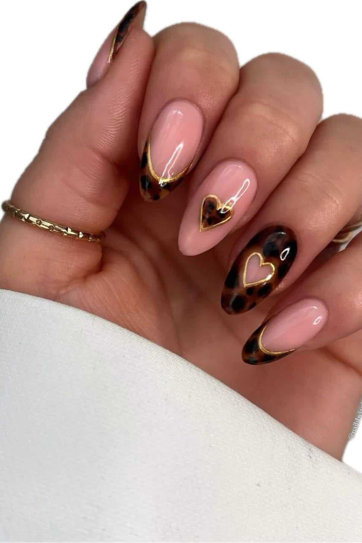 A hand with almond-shaped nails featuring a nude base, leopard print tips, and gold outlined heart designs on two nails.