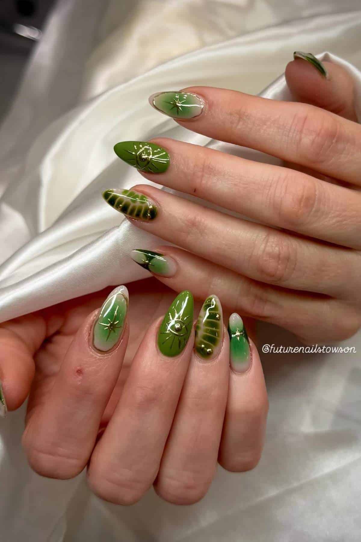 Hands with long almond-shaped nails painted in green and beige with leaf and plant designs, some featuring small gems, on a white satin background.