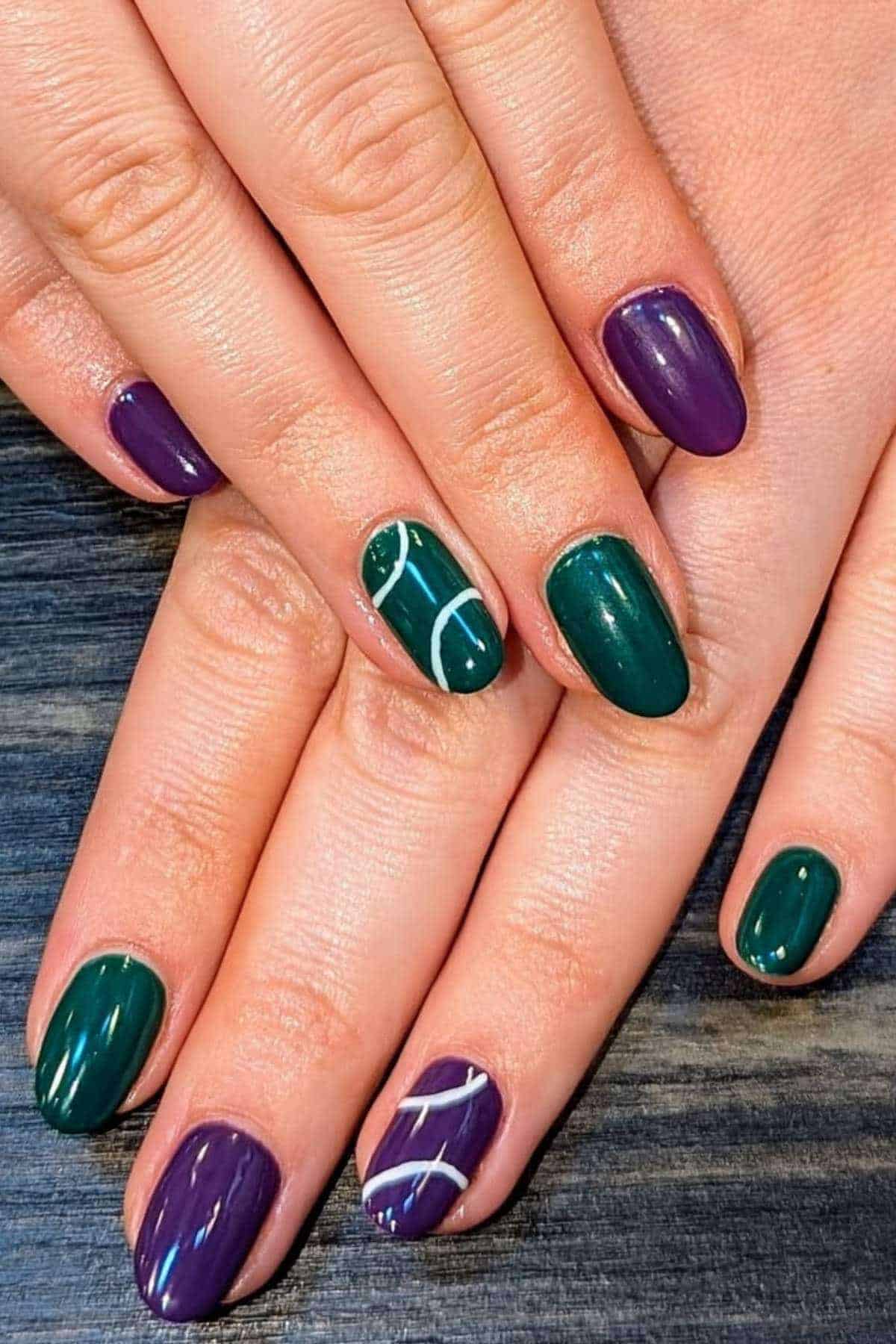 Hands with short nails painted in alternating dark green and purple polish; two nails feature simple white line designs as accents.