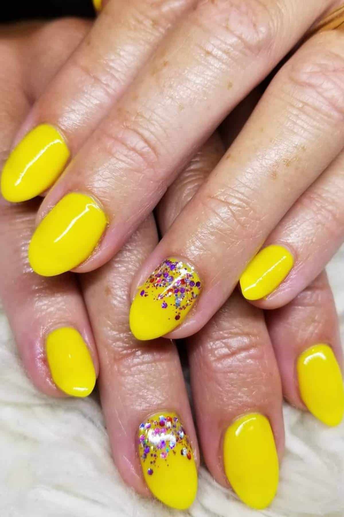 Hands with short, oval-shaped nails painted bright yellow; two nails feature multicolored glitter accents on a yellow base.