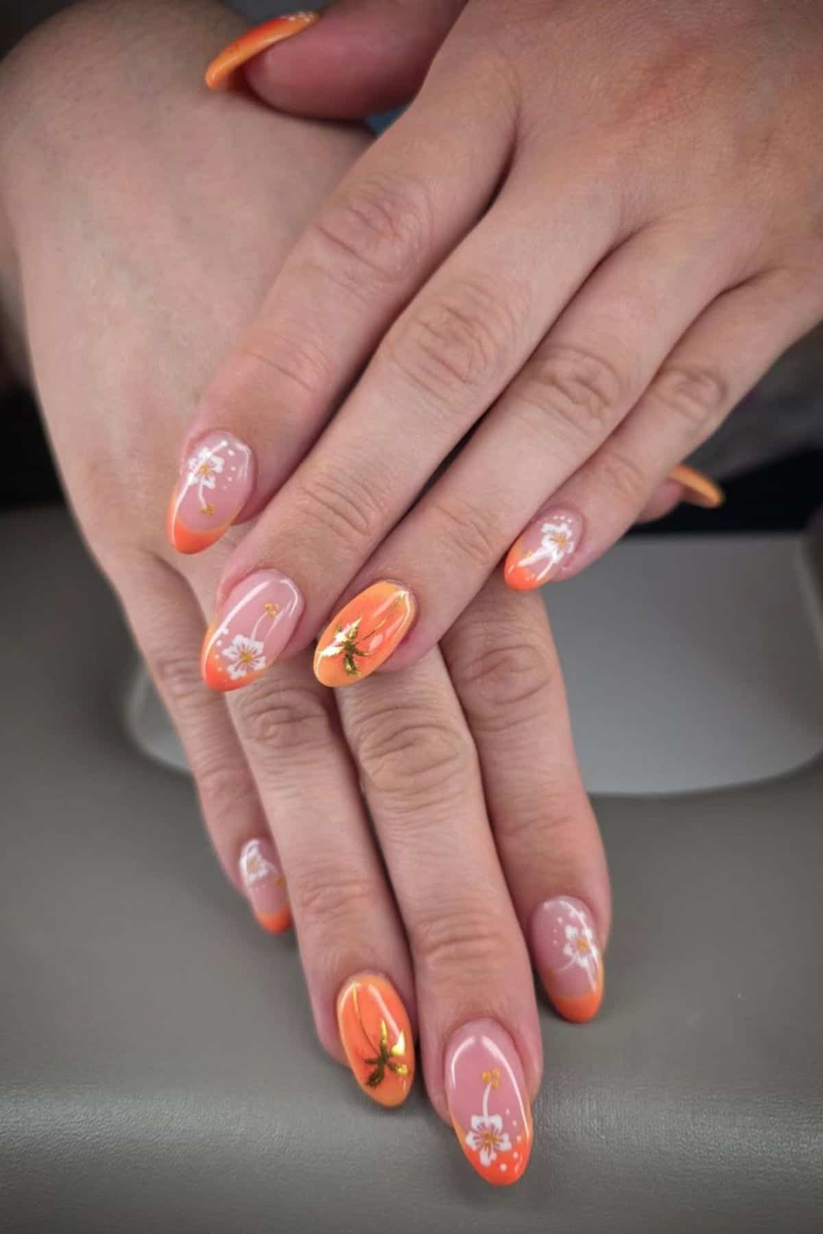 Hands with long, almond-shaped nails painted orange with white floral designs and small gold accents, shown resting on a gray surface.