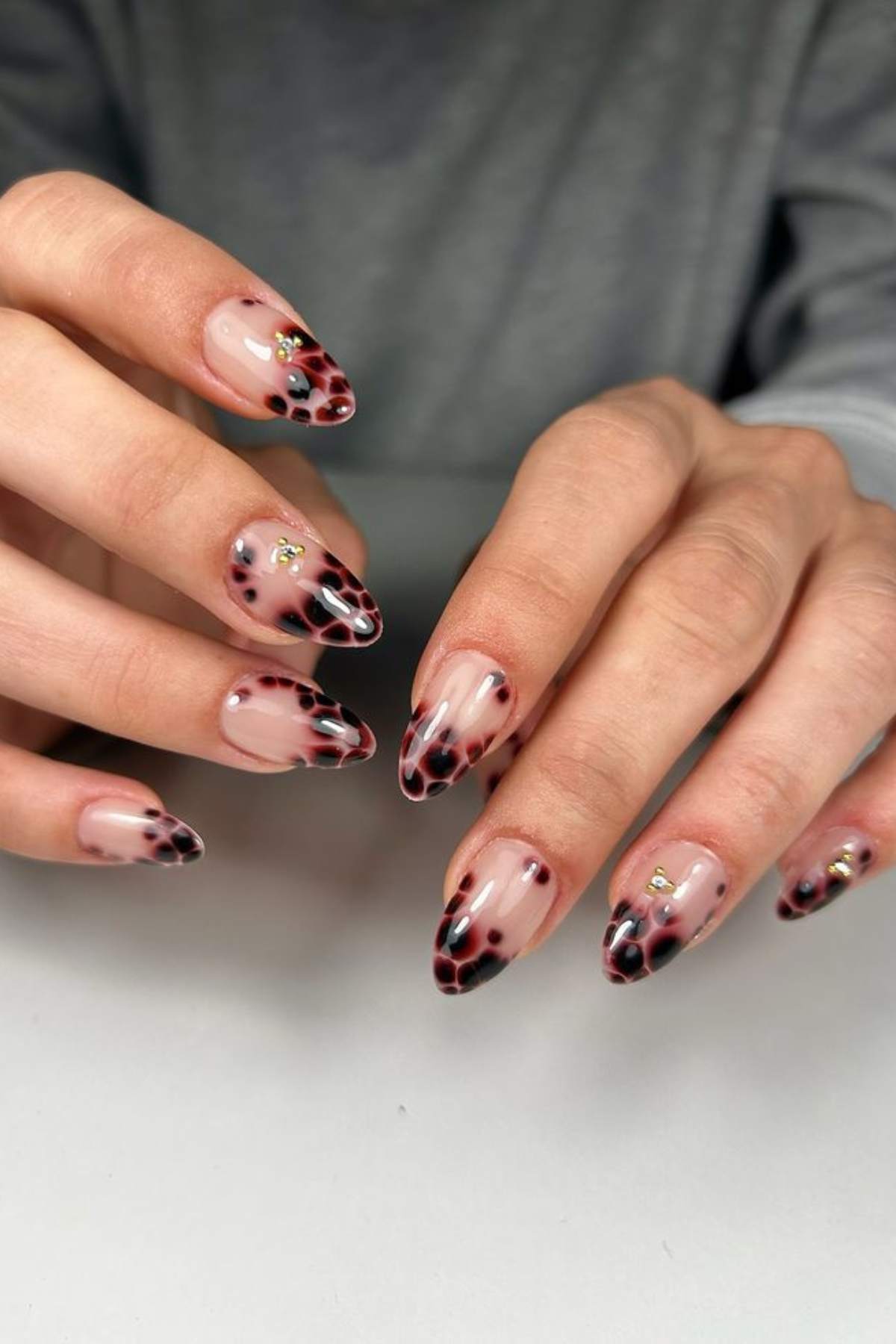 Hands with almond-shaped nails featuring a nude base, black and burgundy leopard print tips, and small gold rhinestone accents on two nails.
