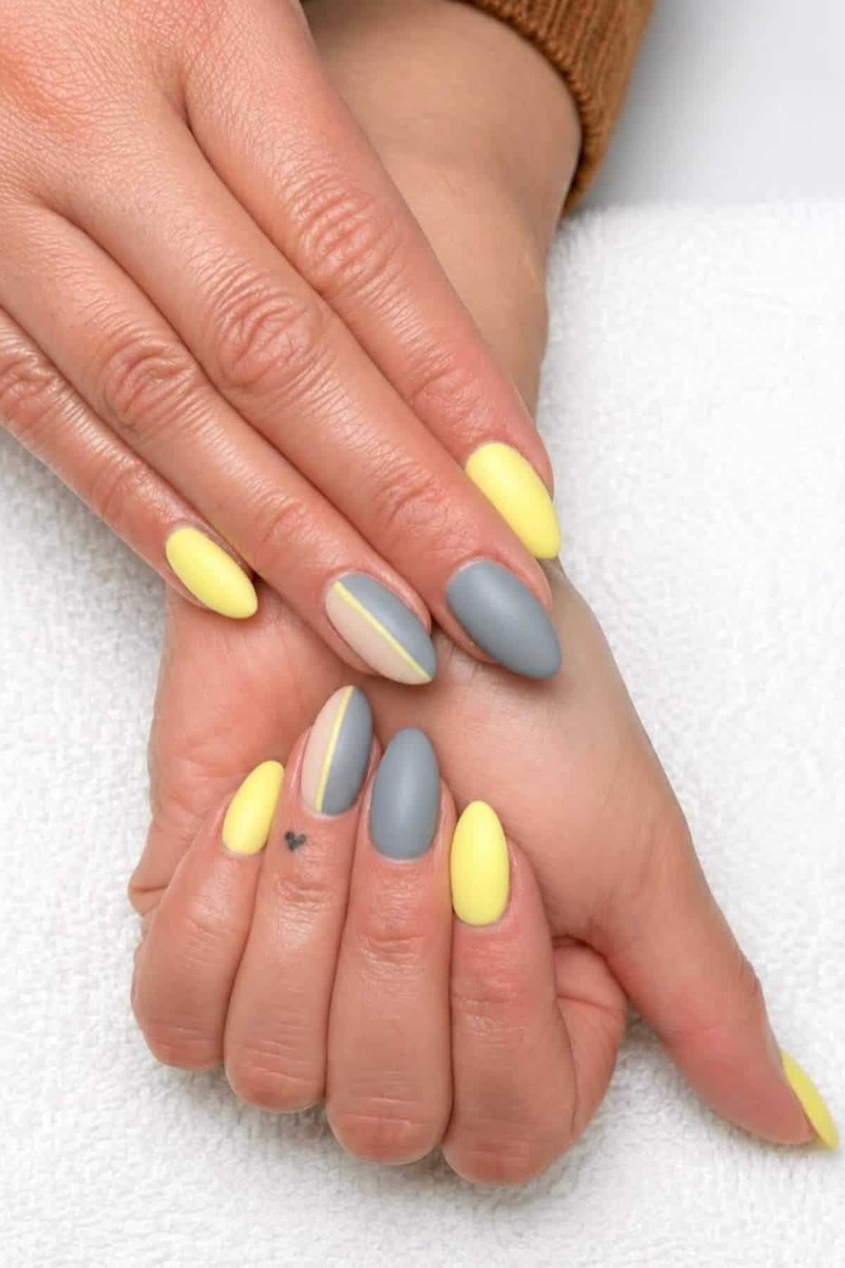 Hands with almond-shaped nails painted in alternating matte yellow and gray, with accent nails featuring yellow and gray stripes. A small heart tattoo is visible on one finger.