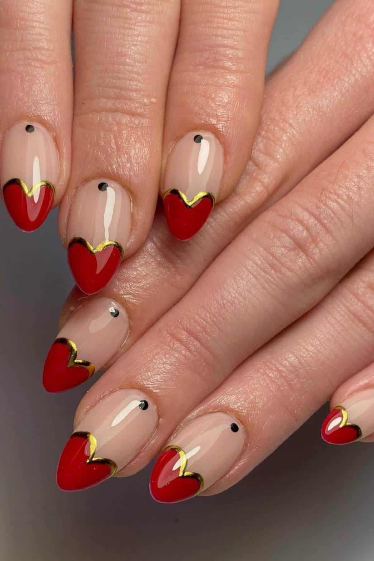 Hands with almond-shaped nails featuring a nude base, red heart-shaped tips, gold outline accents, and small black dots near the cuticles.