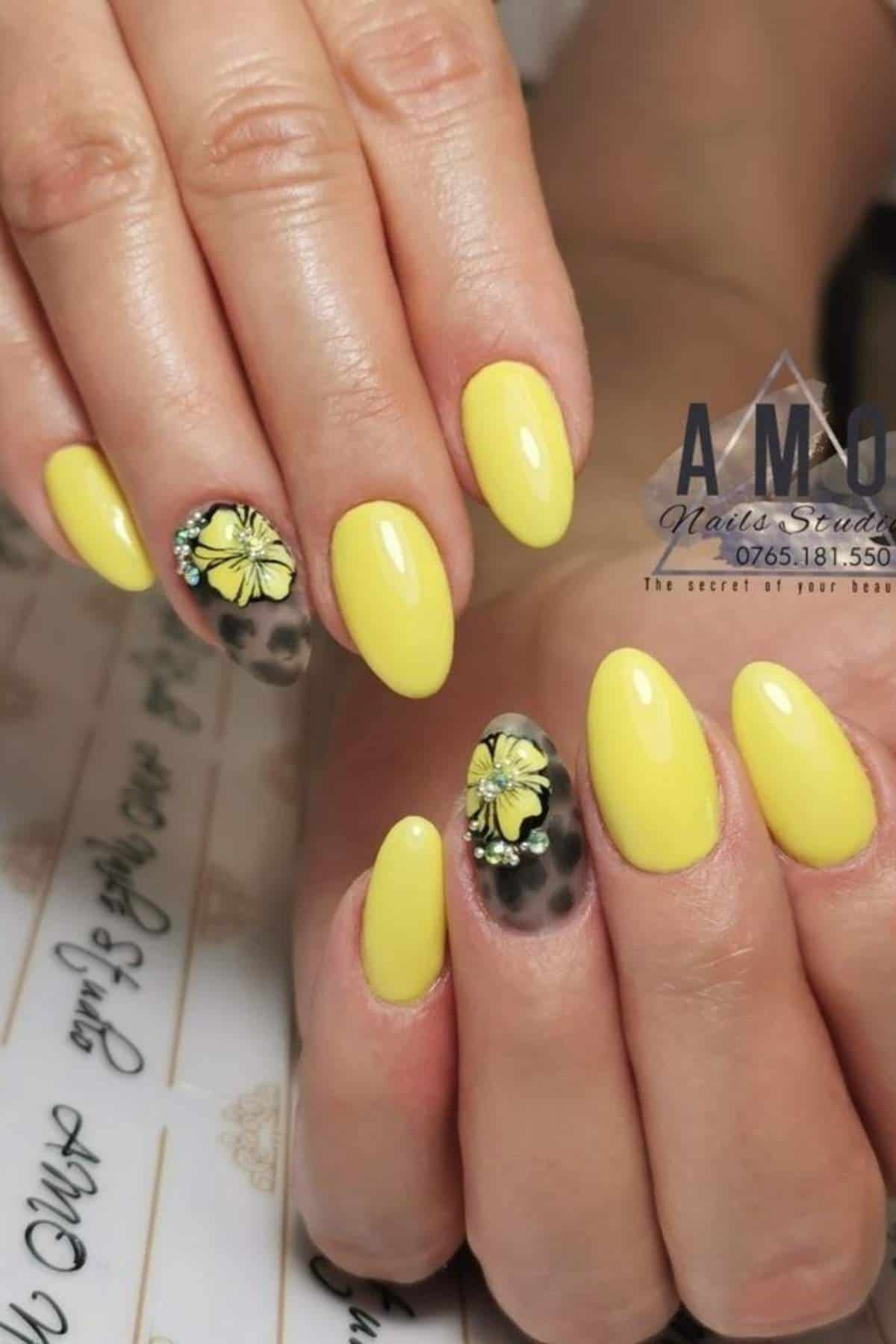 Hands with almond-shaped yellow nails; two accent nails feature a floral design with yellow flowers on a translucent base.