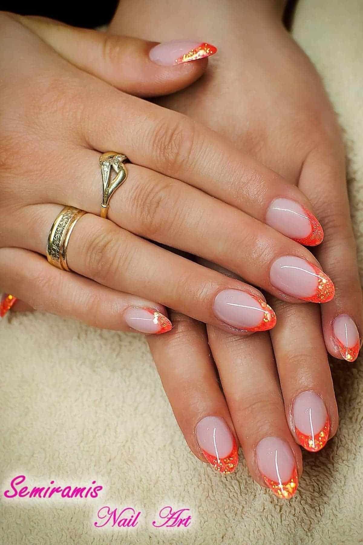 Hands with almond-shaped nails featuring a nude base and orange glitter French tips, resting on a towel. The person is wearing two gold rings.