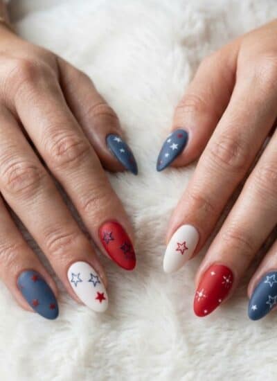 Hands with long almond-shaped nails painted in red, white, and blue with small star designs, resting on a white fuzzy surface.