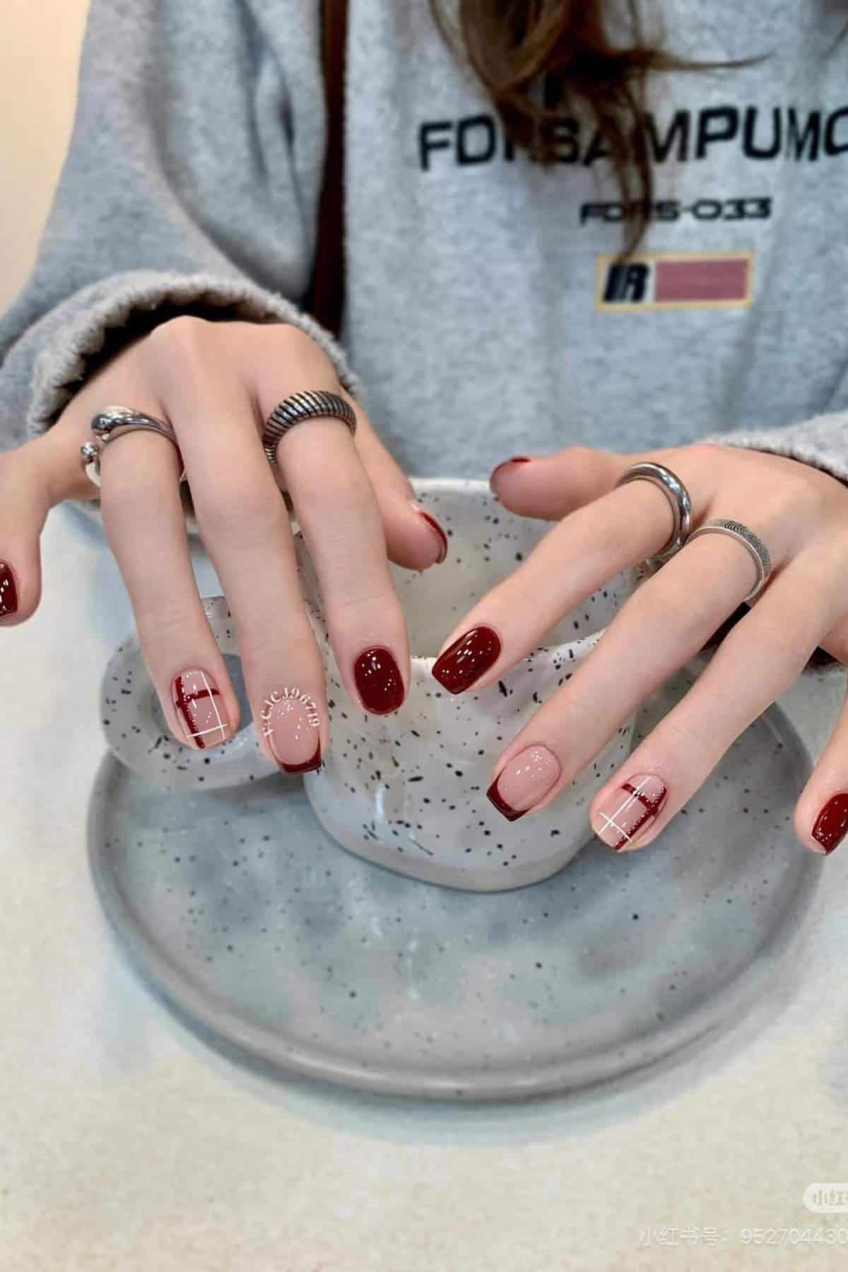 A person with manicured nails featuring red and nude polish, plaid and dot designs, wearing silver rings, holding a speckled ceramic cup over a matching plate.