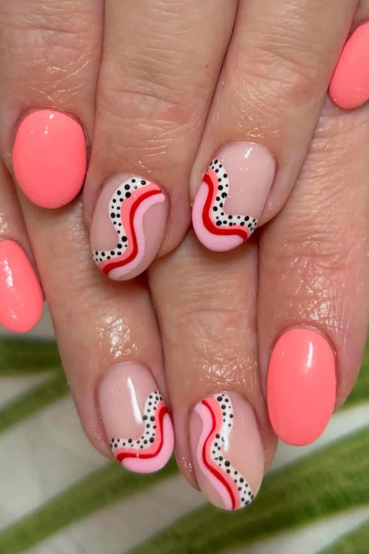 Hands with short nails painted coral pink; some nails feature wavy red and pink lines with black and white dots on a nude base.
