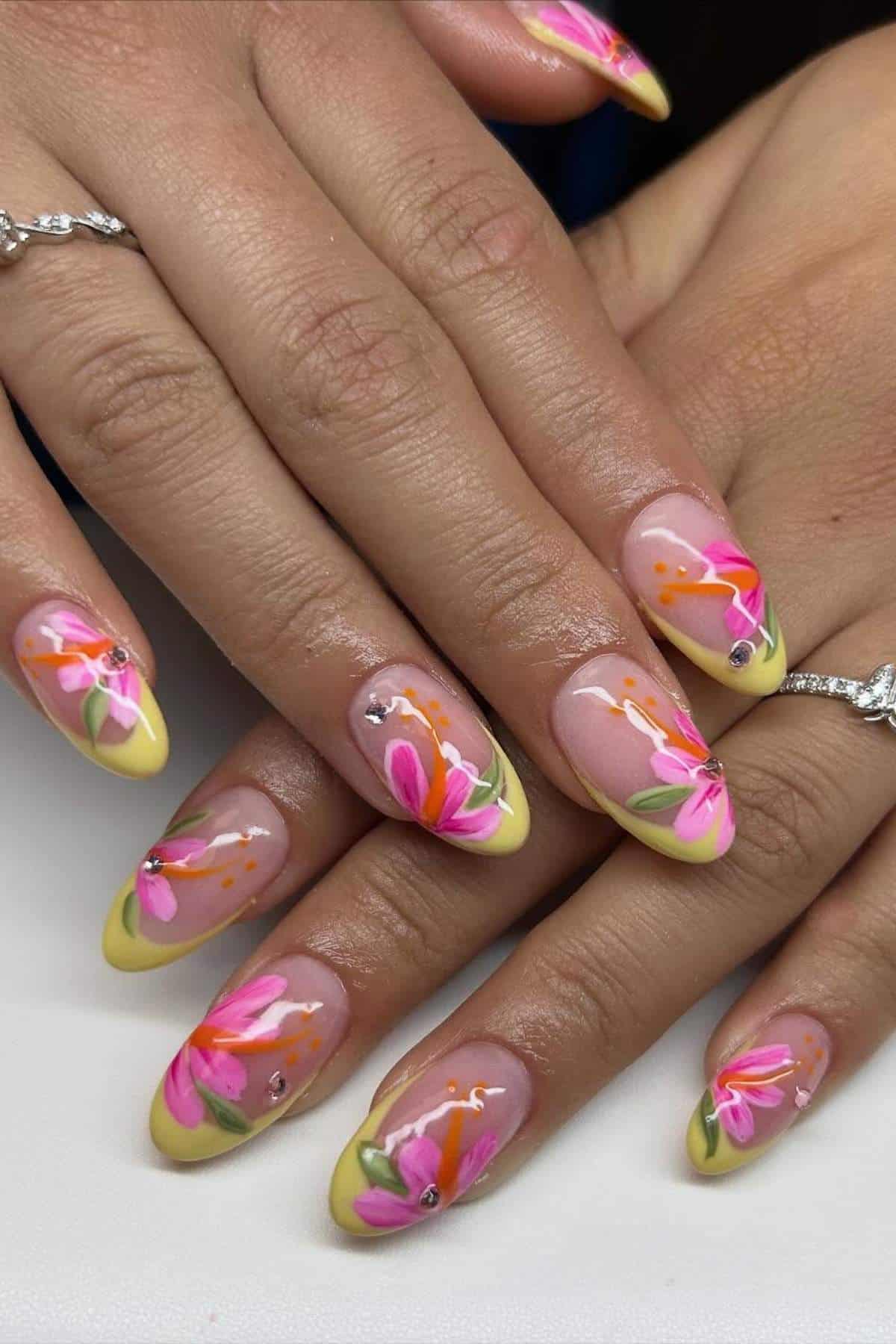 Hands with almond-shaped nails featuring yellow tips, pink and orange floral designs, and small rhinestone accents.
