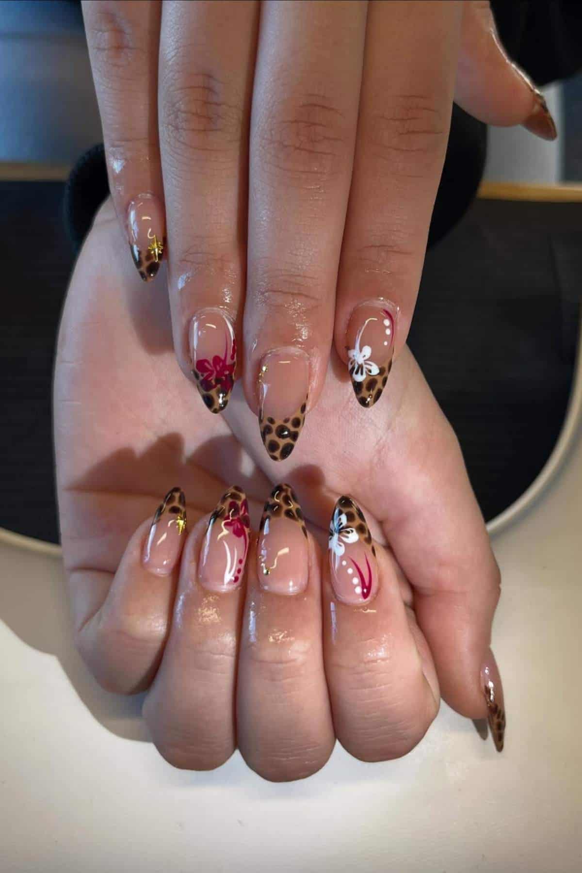 A pair of hands with almond-shaped nails featuring leopard print French tips and colorful floral designs on each nail.