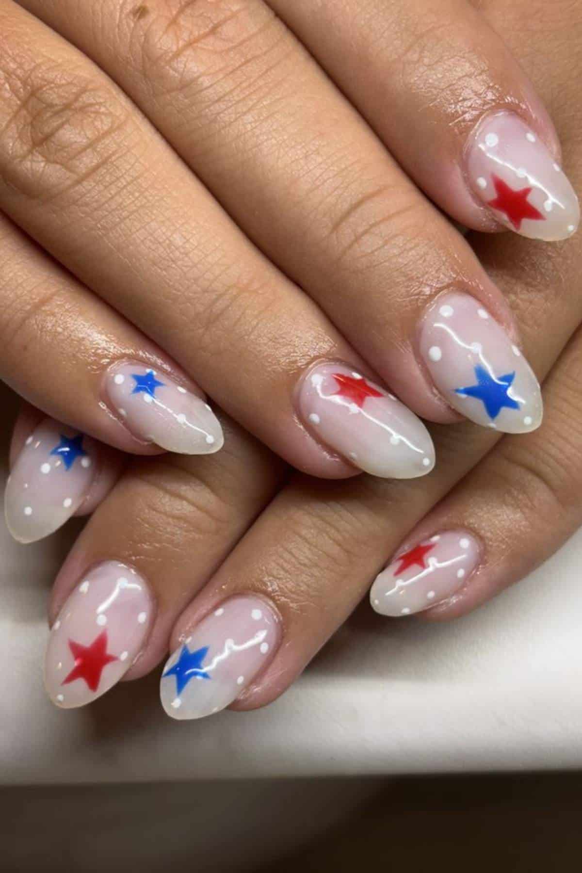 A hand with almond-shaped nails featuring a clear base, white tips, and red and blue star designs, along with small white dots.