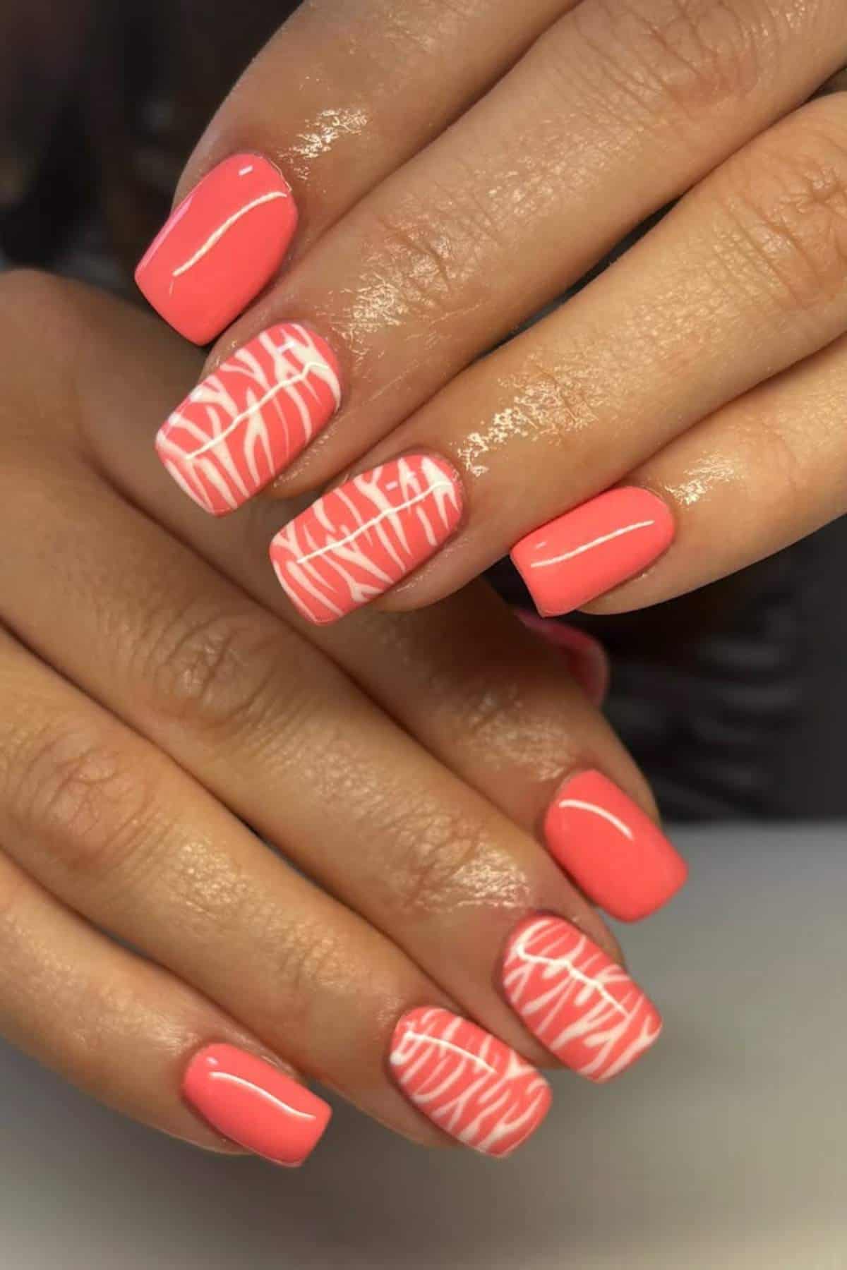 Hands with short, square-shaped coral pink nails; some nails feature white abstract line designs on the same pink base.