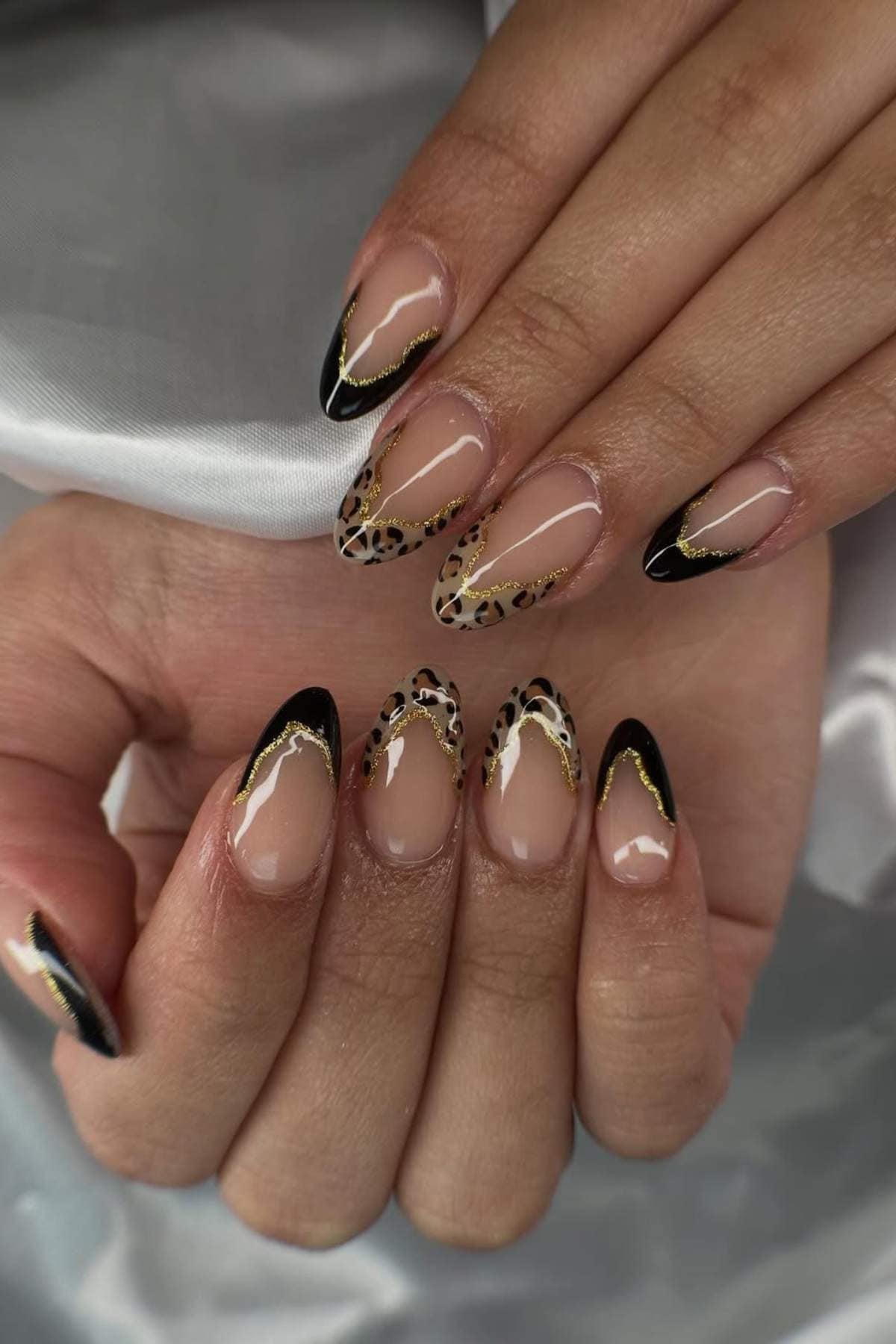 Hands with almond-shaped nails featuring a nude base, black and gold tips, and leopard print accents on some nails.