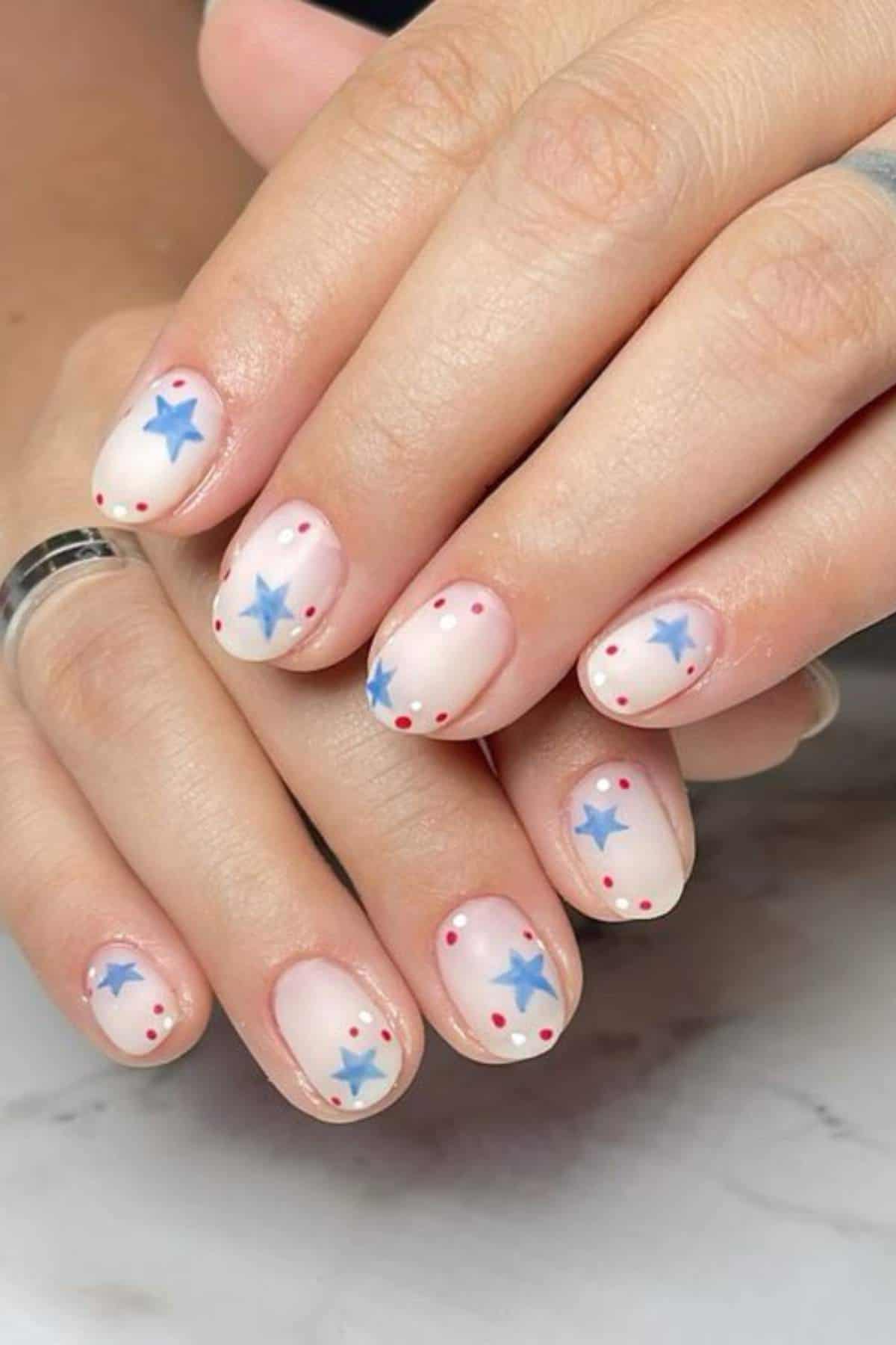 Close-up of hands with short nails painted in a light pink base, decorated with blue stars and small red dots on each nail.
