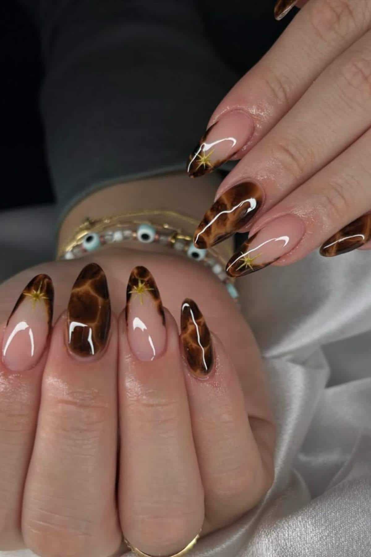 Close-up of hands with almond-shaped nails featuring a brown marble design with gold accents and a natural pink base.