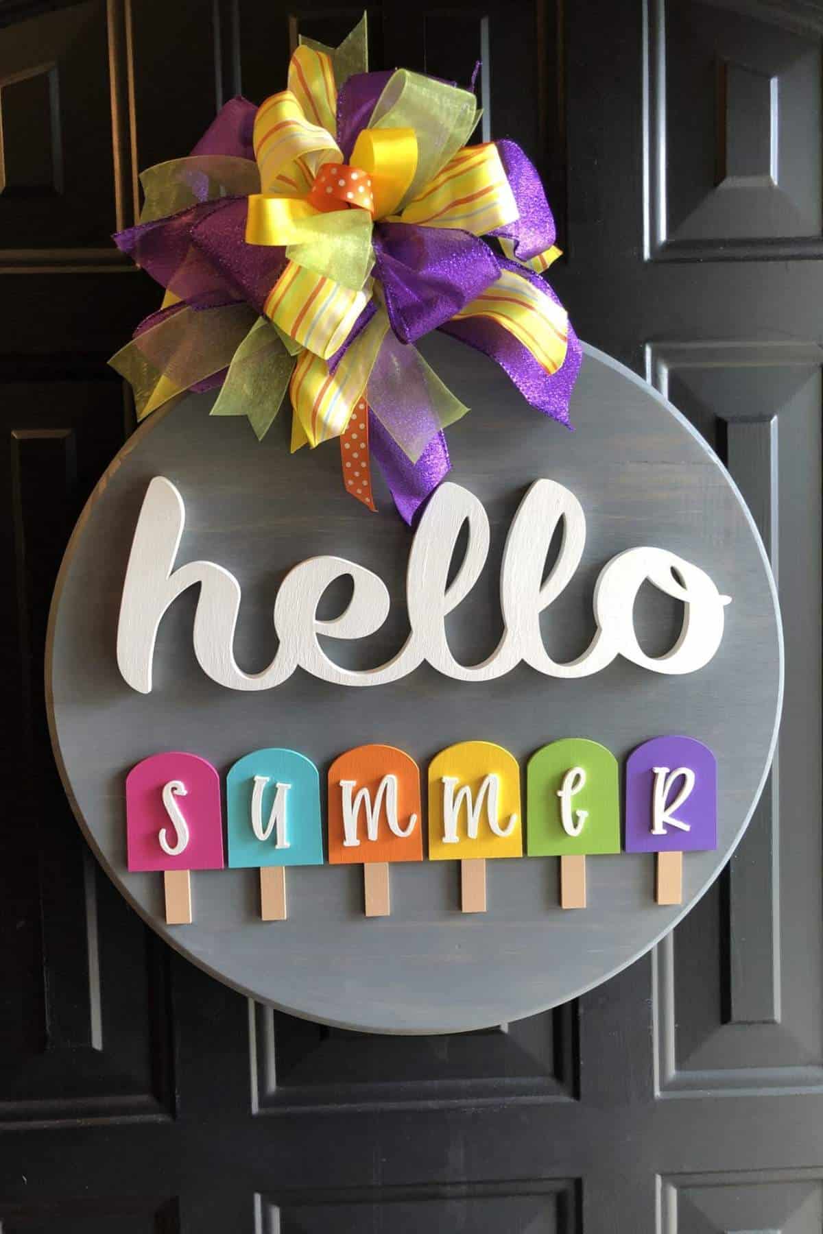 A round sign with a colorful bow says "hello summer" with the word "summer" spelled on popsicle shapes in bright colors.