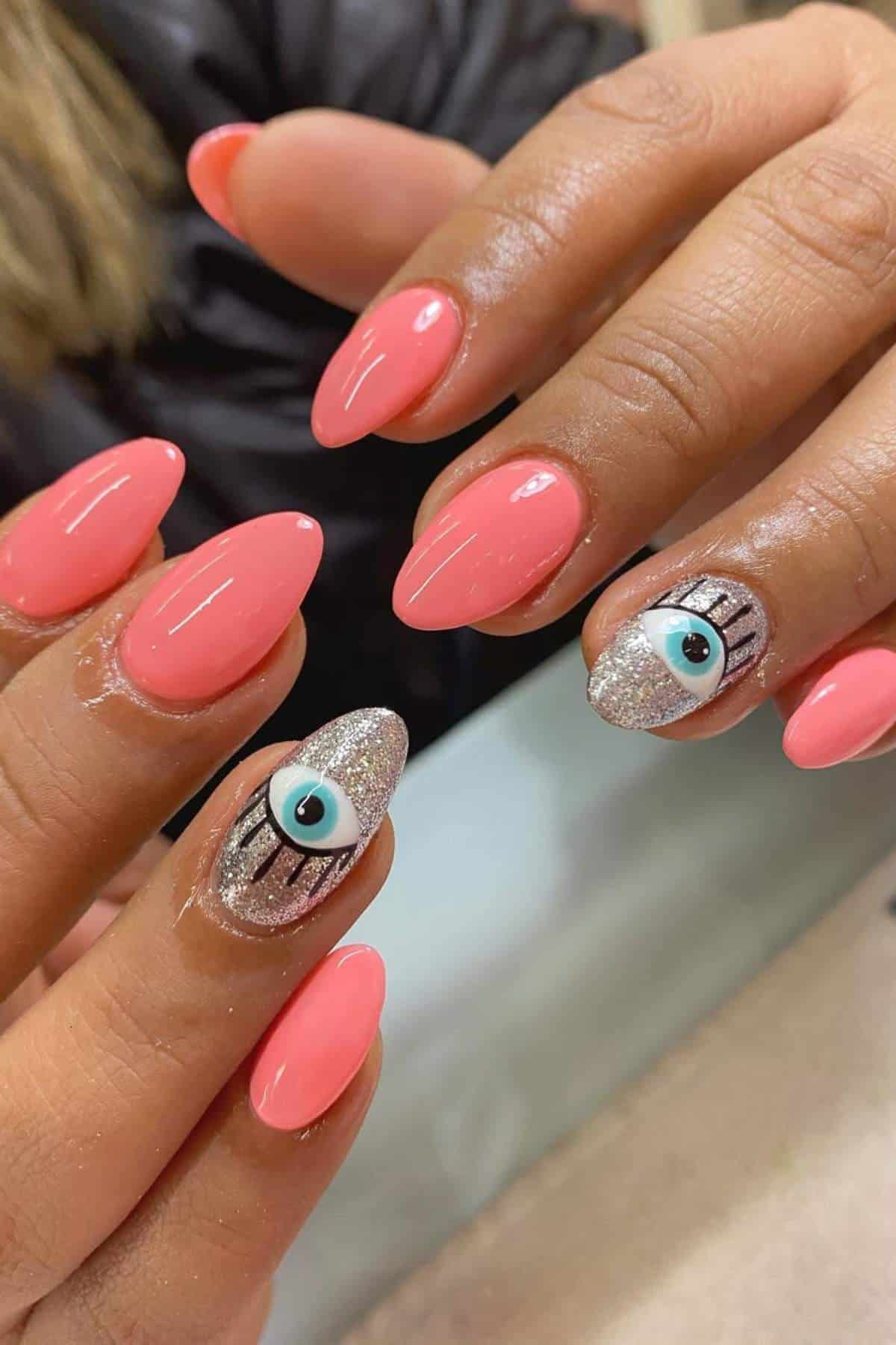 Hands with glossy, almond-shaped coral nails; ring fingers feature silver glitter polish with an eye design and black eyelash details.