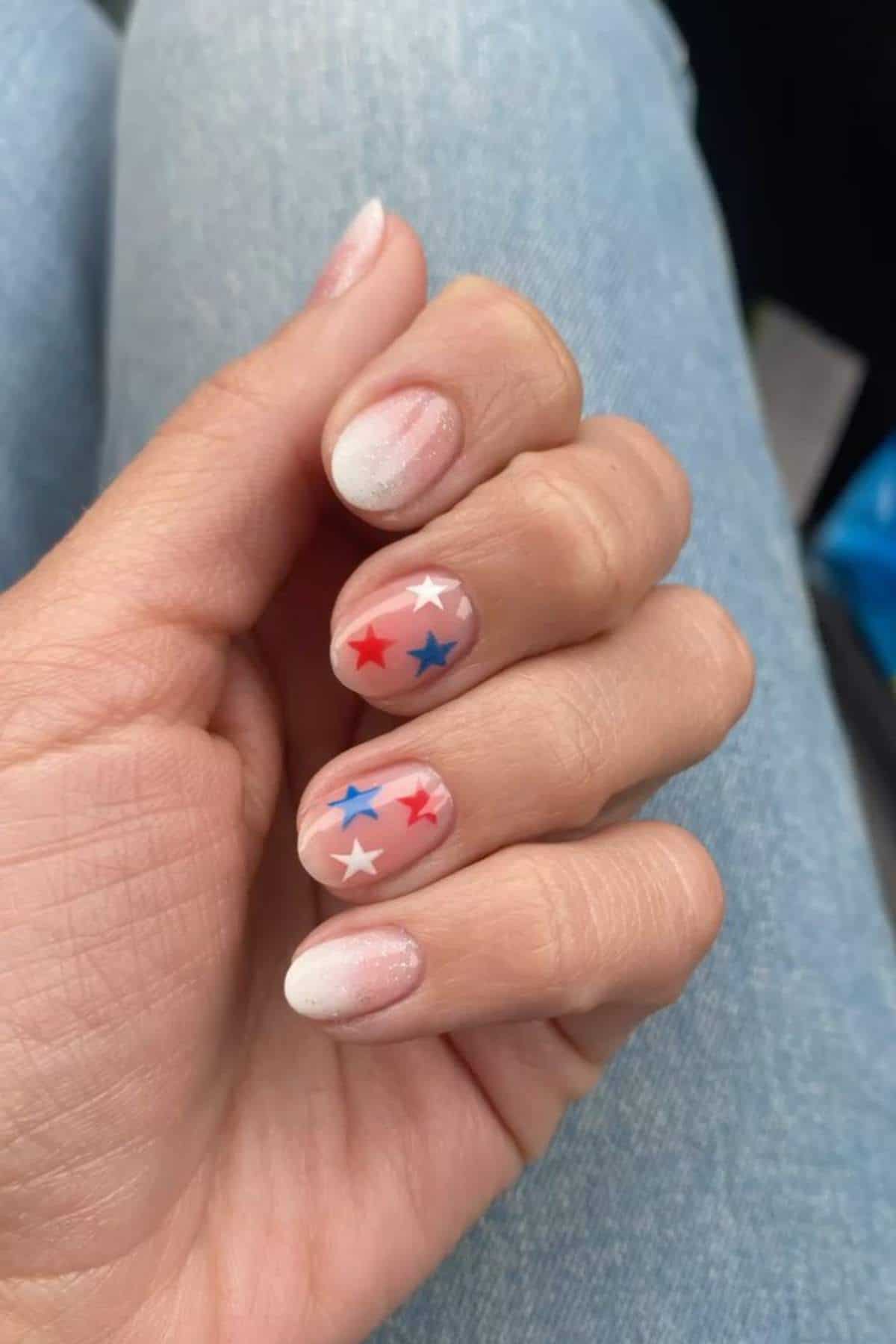 A hand with short, almond-shaped nails featuring a pale gradient base and red, white, and blue star decals on two fingernails. The person is wearing light blue jeans.
