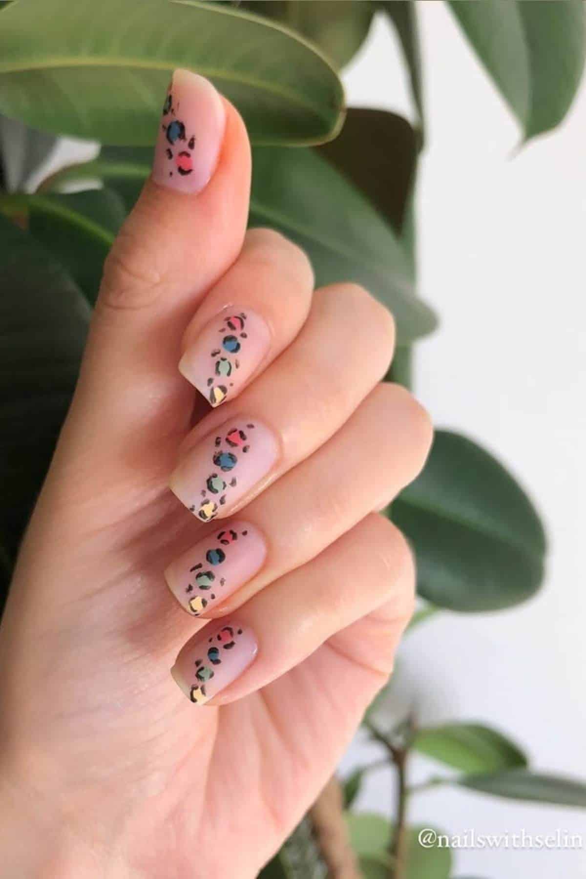 A hand with medium-length square nails featuring a nude base and multicolored leopard print nail art, held in front of green leaves.