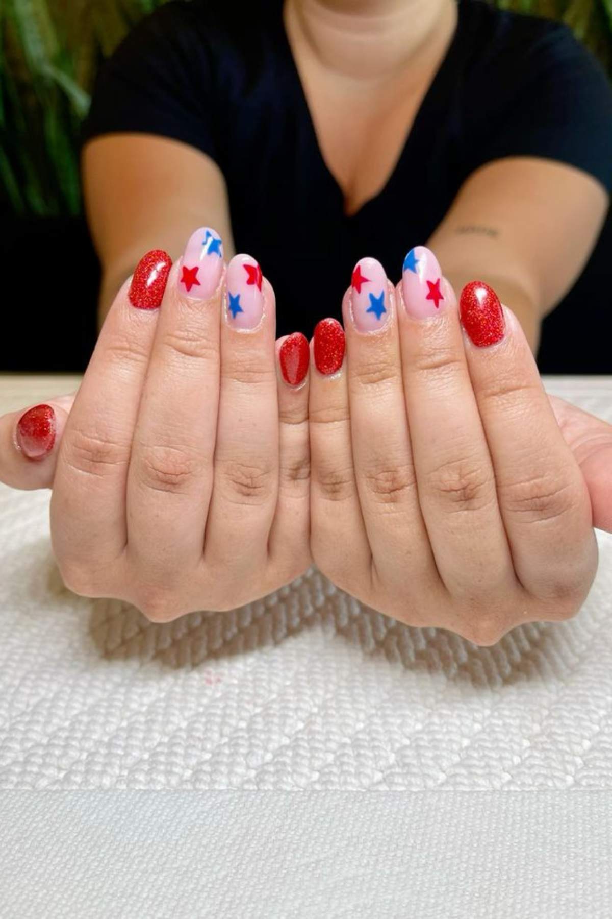 A person shows manicured hands with red glitter polish and white nails featuring red and blue star designs, resting on a textured white surface.