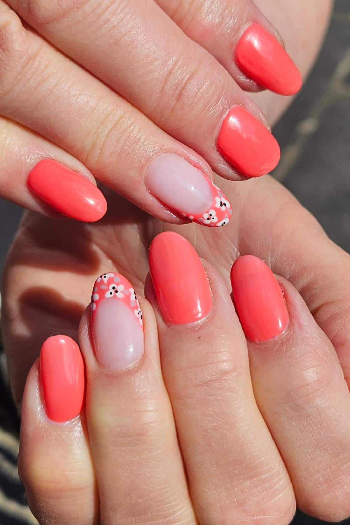Hands with oval-shaped nails painted coral pink; two accent nails have a clear base with small white and pink floral designs near the tips.