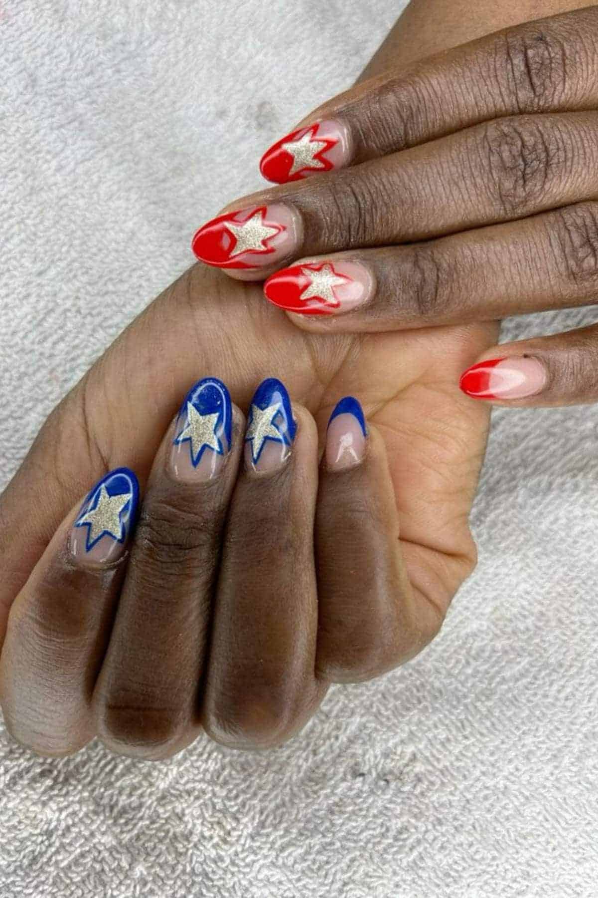 Hands with manicured nails featuring red polish with silver stars on one hand and blue polish with silver stars on the other, resting on a textured white surface.
