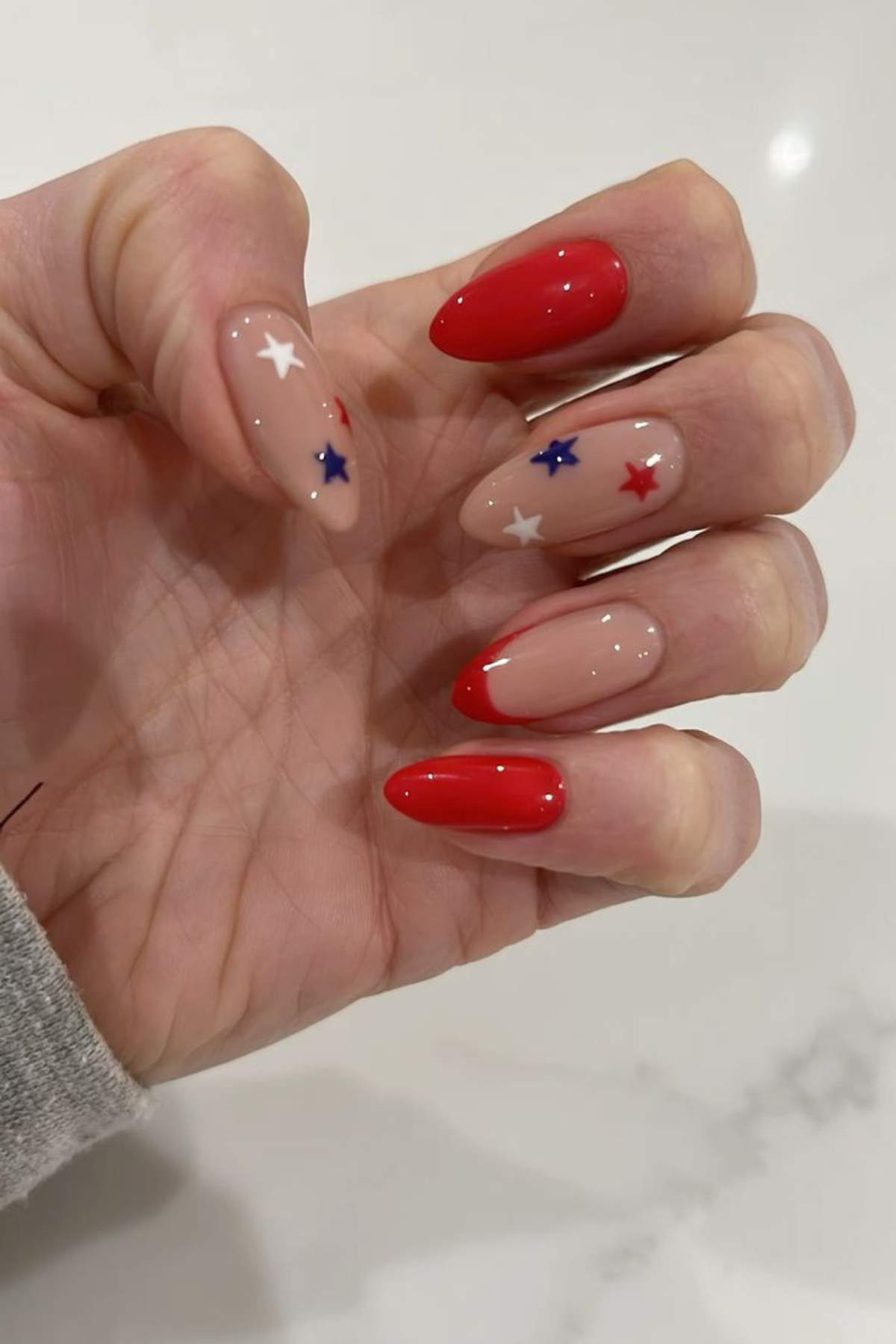 A hand with almond-shaped nails; some are painted red, while others are nude with red and blue star decals and partial red tips.