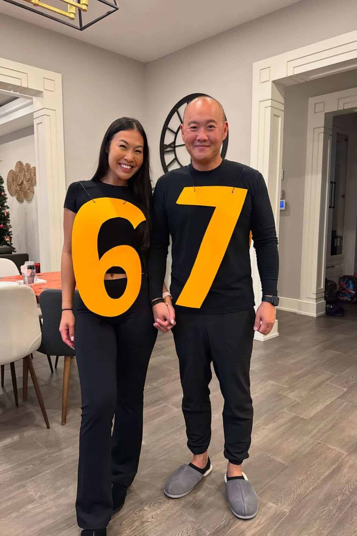 Two people stand indoors holding hands, each wearing a large yellow number; the woman wears a "6" and the man wears a "7." Both are dressed in black.