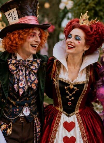 Four people dressed as Alice in Wonderland characters—Alice, the Mad Hatter, the Queen of Hearts, and the Cheshire Cat—stand together smiling at a whimsical, tea party-themed setting.