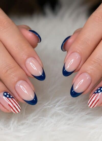 Close-up of hands with almond-shaped nails featuring a blue French tip design and American flag accents on the ring fingers. The hands wear gold rings, one with a diamond.