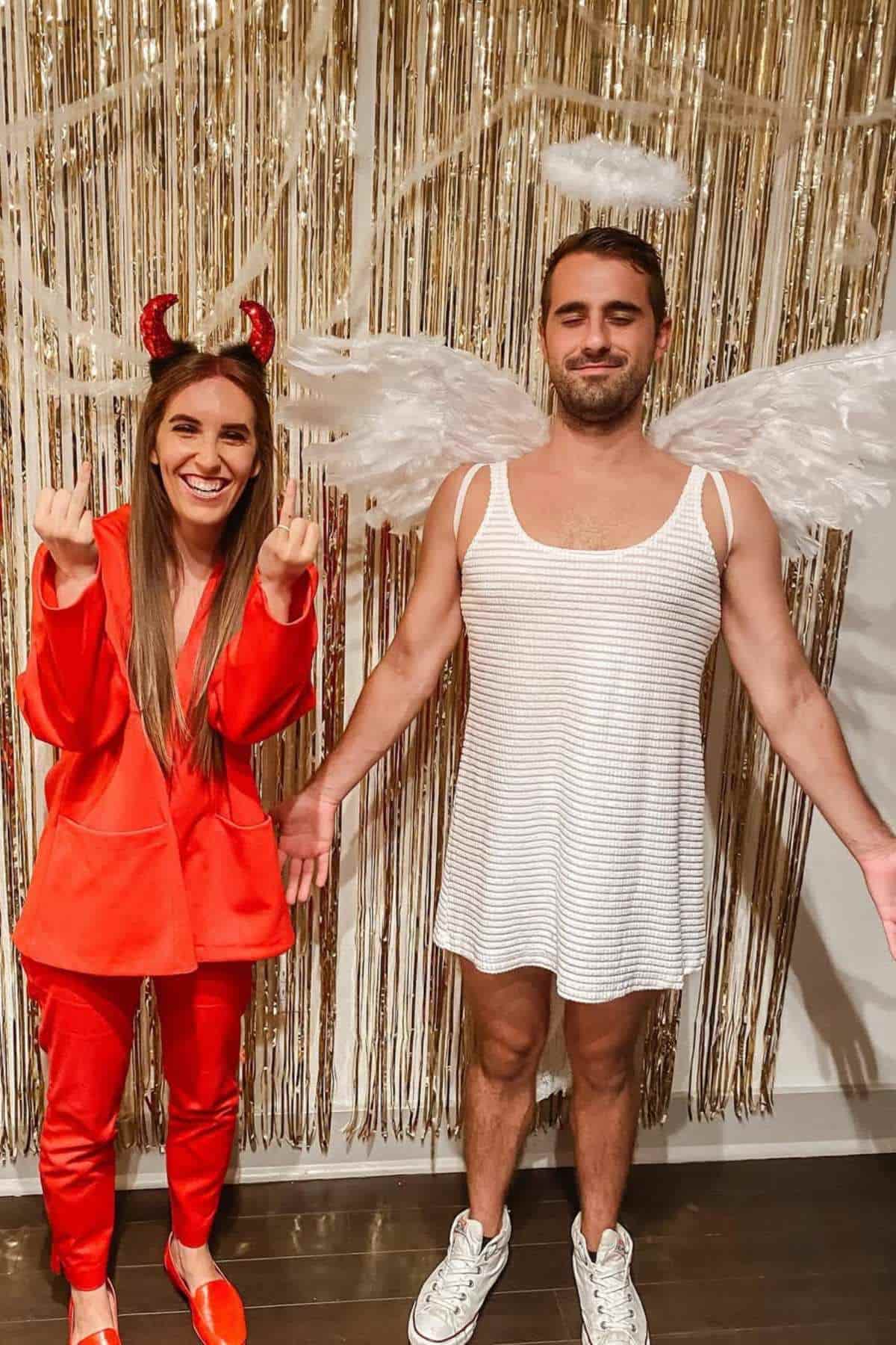 Two people in costumes stand in front of a gold tinsel backdrop. One is dressed as a devil in red, and the other as an angel with wings, a halo, and a white dress.