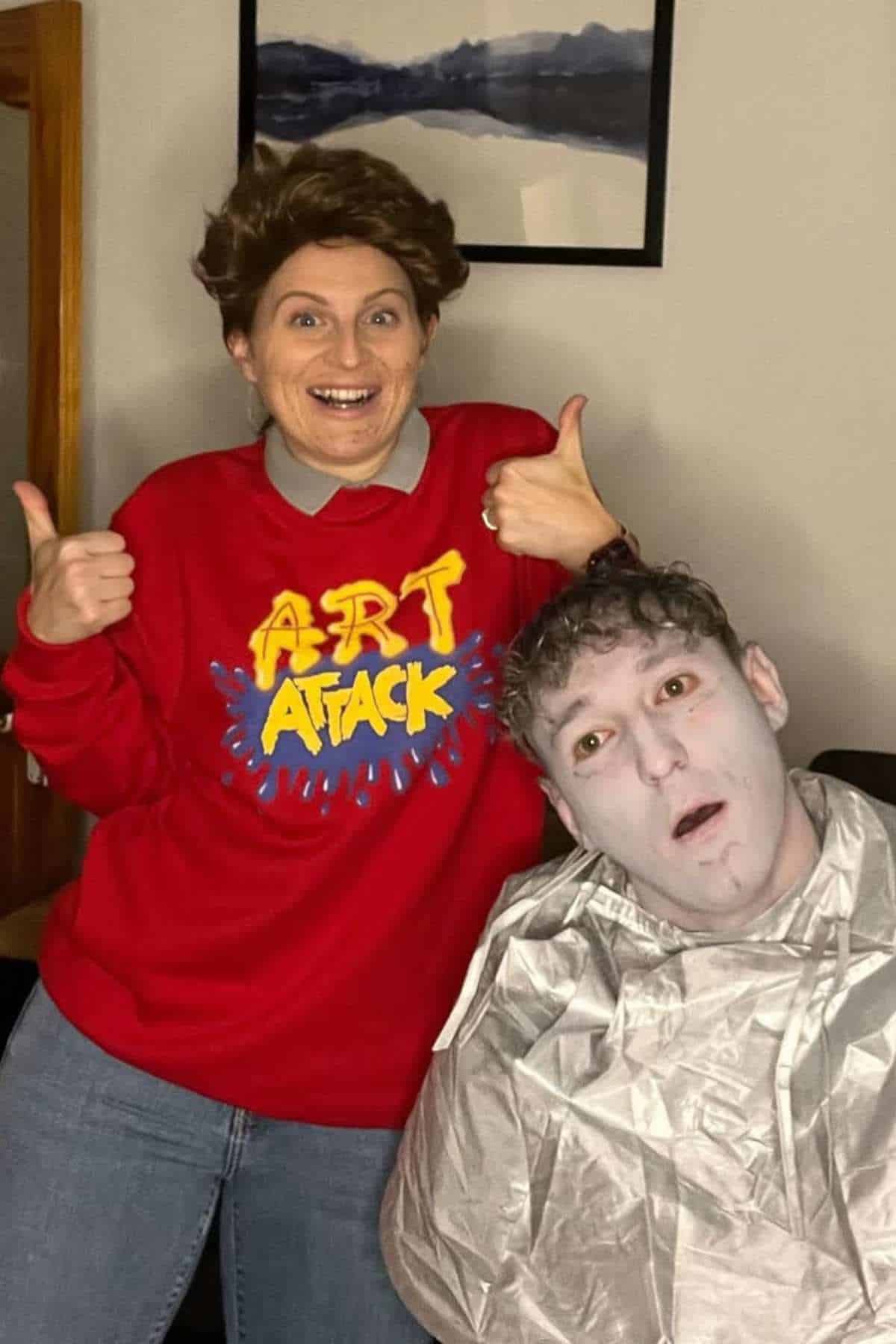 Two people in costume; one in a red "Art Attack" sweater with a wig and thumbs up, the other with face painted pale and wearing a silver cover, sitting down.