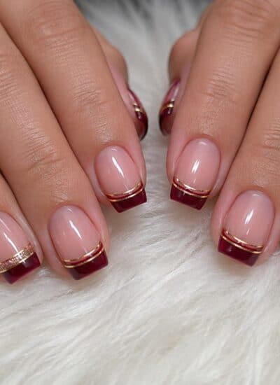 Hands with a manicure featuring deep red French tips and thin gold lines on a light pink base, resting on a white fluffy surface.