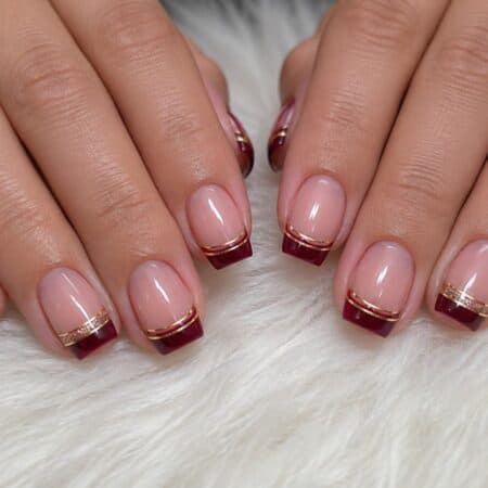 Hands with a manicure featuring deep red French tips and thin gold lines on a light pink base, resting on a white fluffy surface.