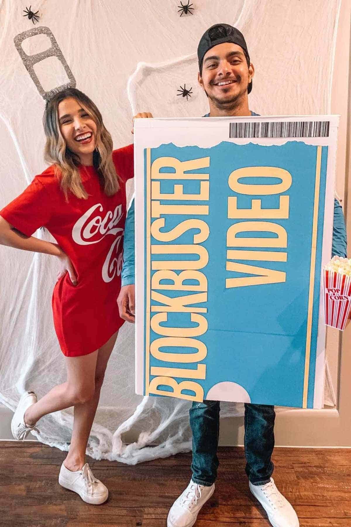 A woman in a Coca-Cola dress stands next to a man wearing a large Blockbuster Video box costume, both smiling at the camera.
