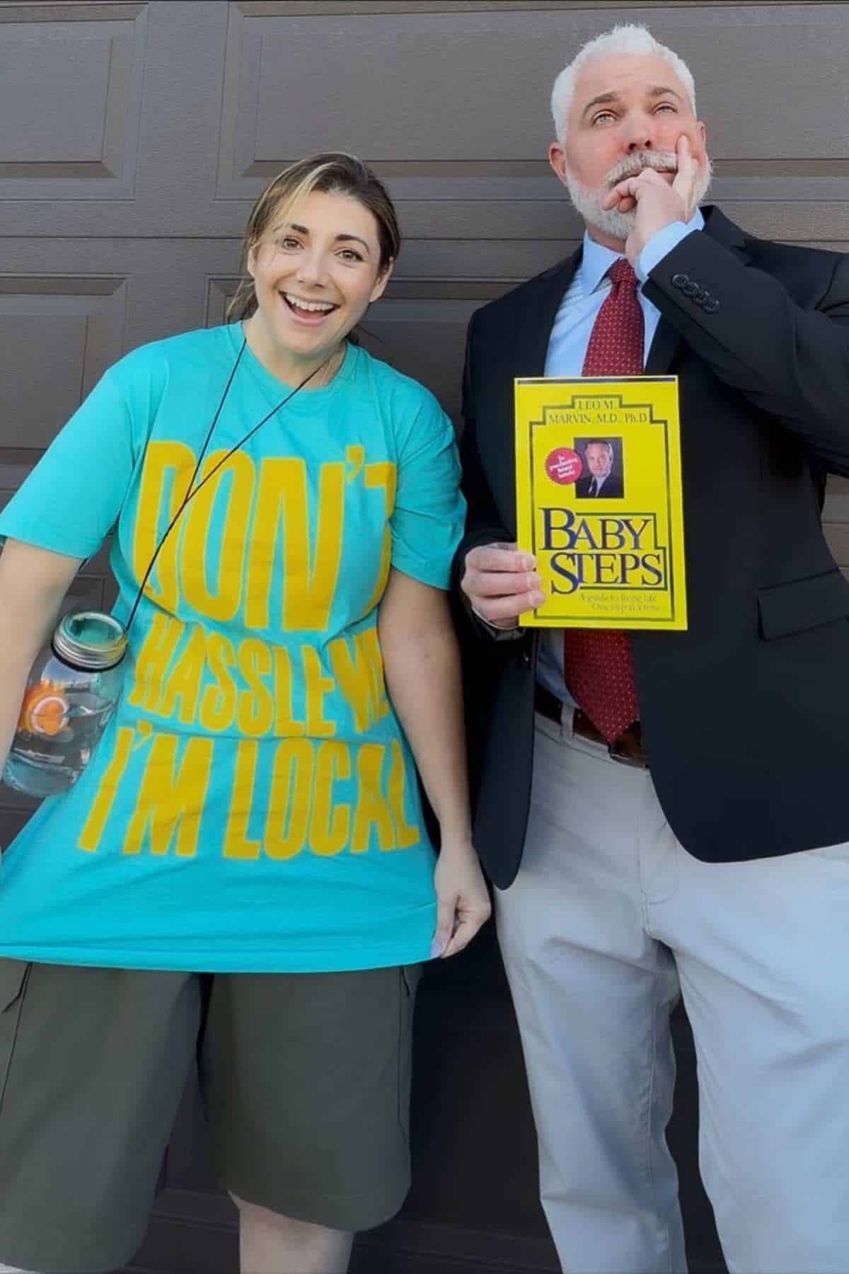 Two people stand in front of a garage; one wears a teal shirt that says "Don't hassle me I'm local," and the other holds a "Baby Steps" book and bites their finger.