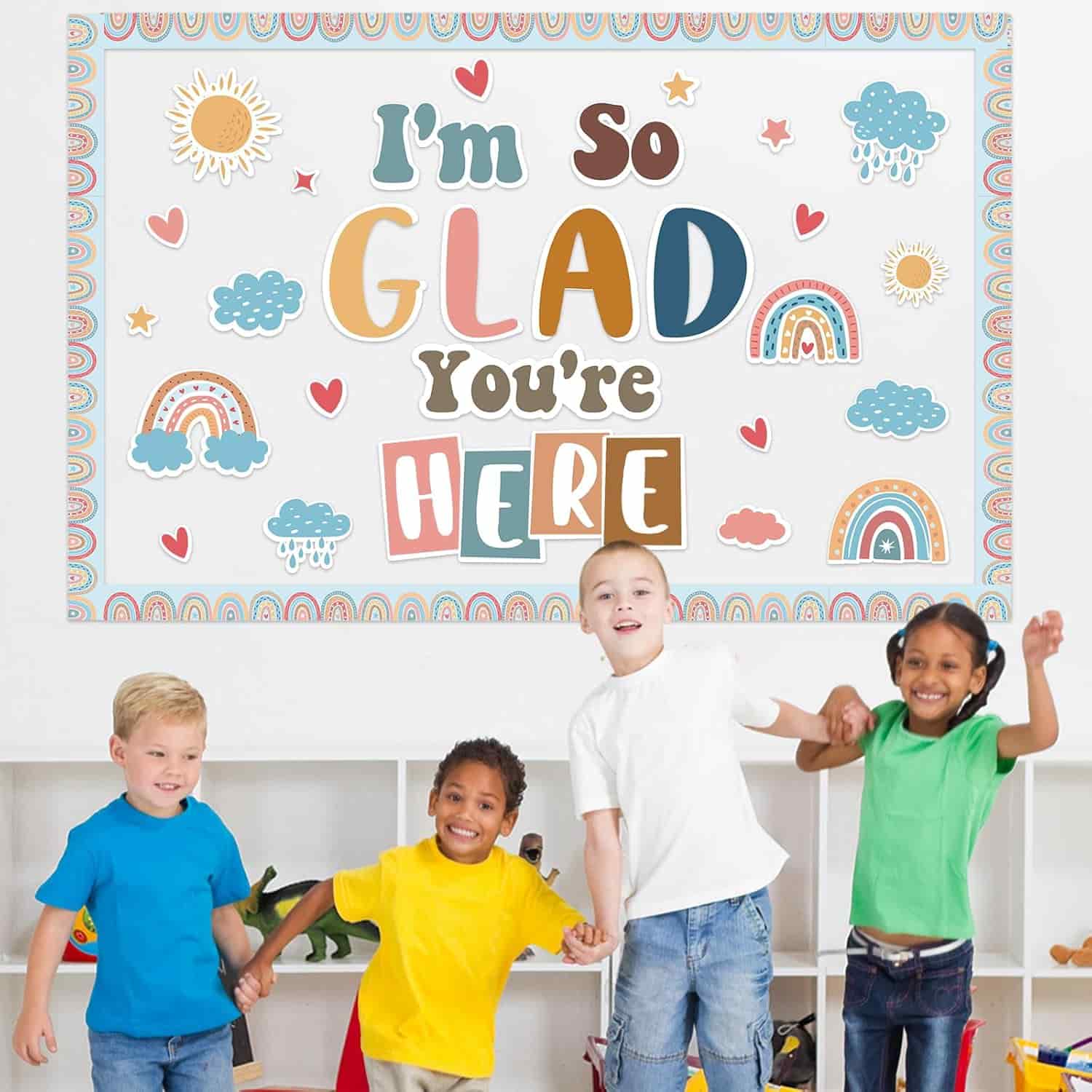 Four young children stand smiling and holding hands in front of a colorful sign that reads, "I'm so glad you're here," with rainbow and cloud illustrations.