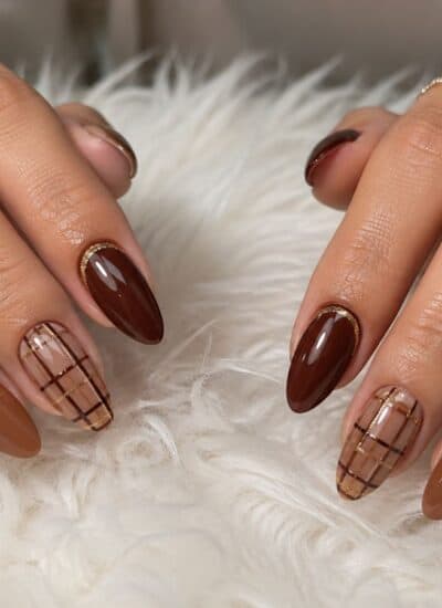 Hands with almond-shaped nails featuring brown and nude tones, some with plaid designs, resting on a white fluffy surface.
