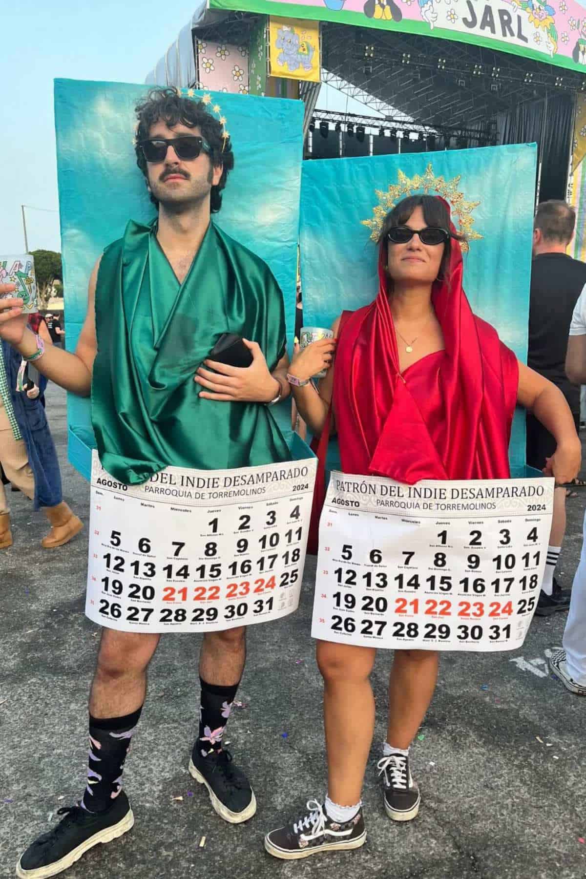 Two people stand outdoors wearing costumes made of green and red fabric with large calendar pages for August 2024, each referencing the patron saint festival of Torremolinos.