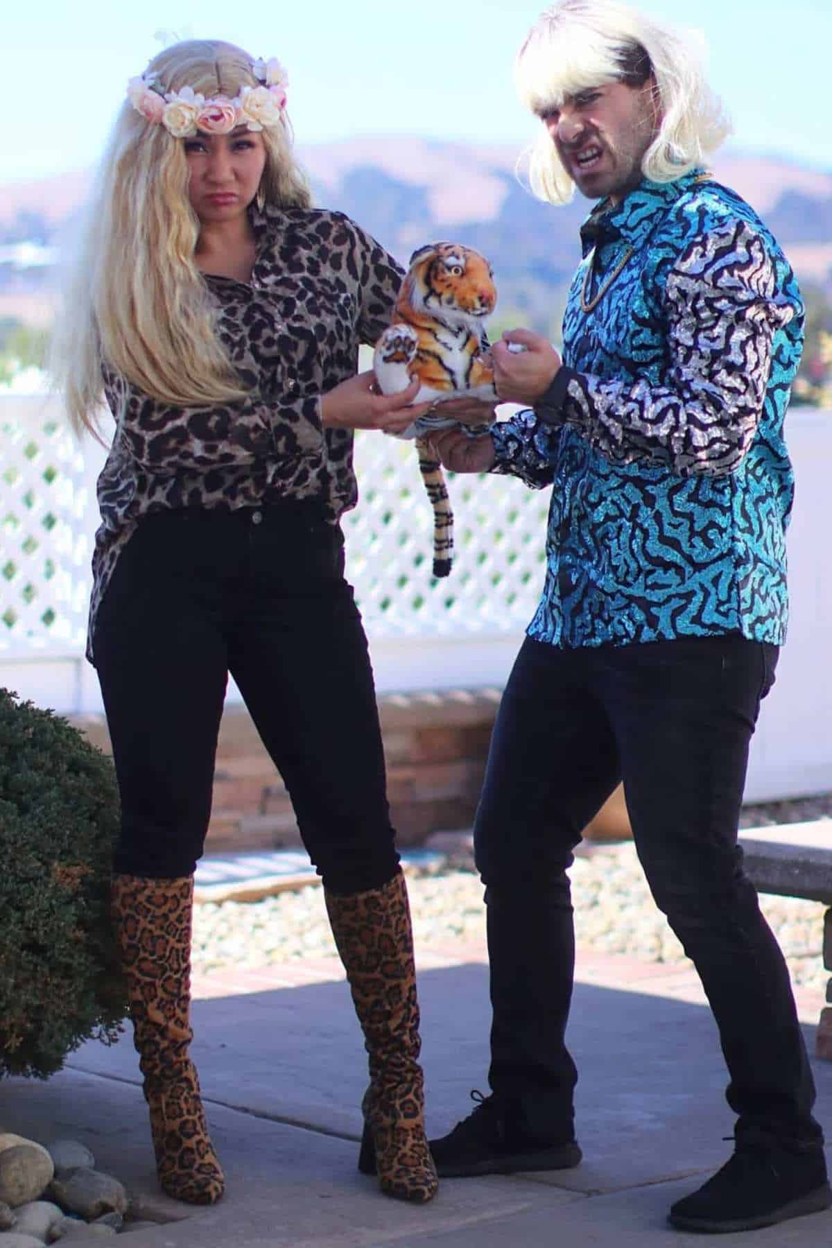 Two adults wearing animal print clothing and blonde wigs hold a tiger plush toy outside, with mountains visible in the background.