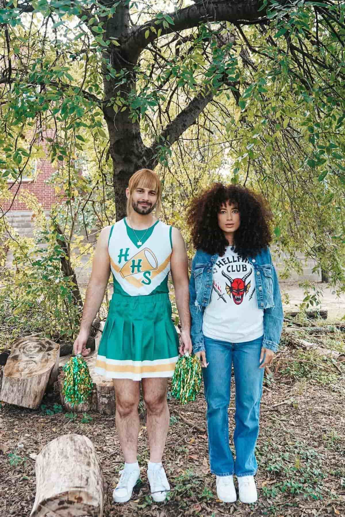 Two people stand outdoors; one wears a green cheerleader uniform with pom-poms, the other wears a denim jacket, graphic T-shirt, and jeans.