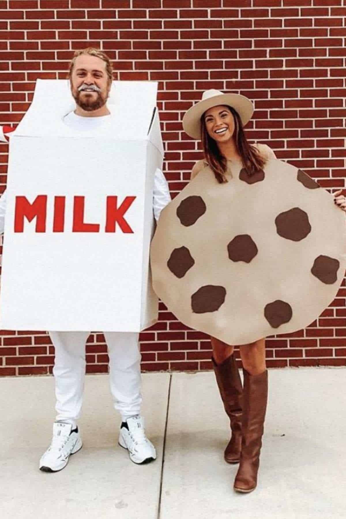 Two people stand in front of a brick wall; one wears a milk carton costume, the other wears a chocolate chip cookie costume and a hat.