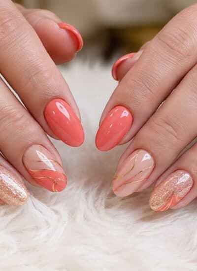 Hands with manicured nails painted in coral, gold glitter, and nude shades, featuring gold line designs, resting on a white fluffy surface.