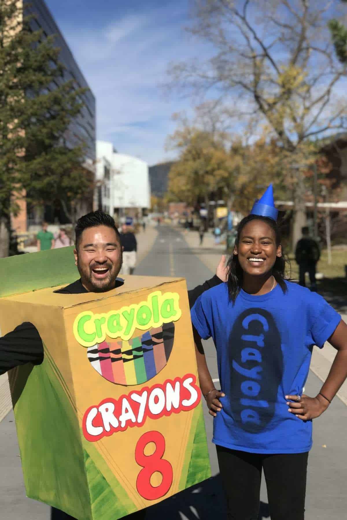 Two people stand outdoors in costume; one is dressed as a Crayola crayon box, and the other is wearing a blue shirt and cone hat resembling a blue crayon.