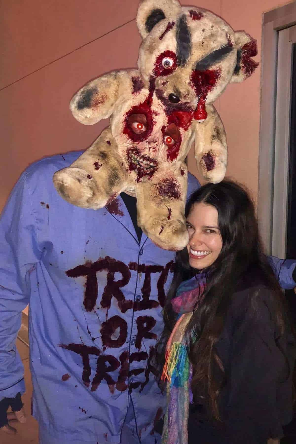 A person wearing a bloody, zombie teddy bear mask and a shirt that says "Trick or Treat" stands next to a smiling woman in a colorful scarf.